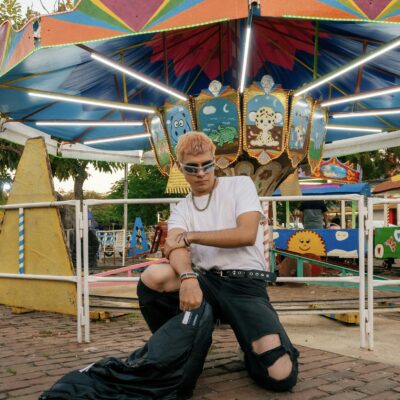 A stylish young man models in front of a colorful carousel at a lively amusement park.