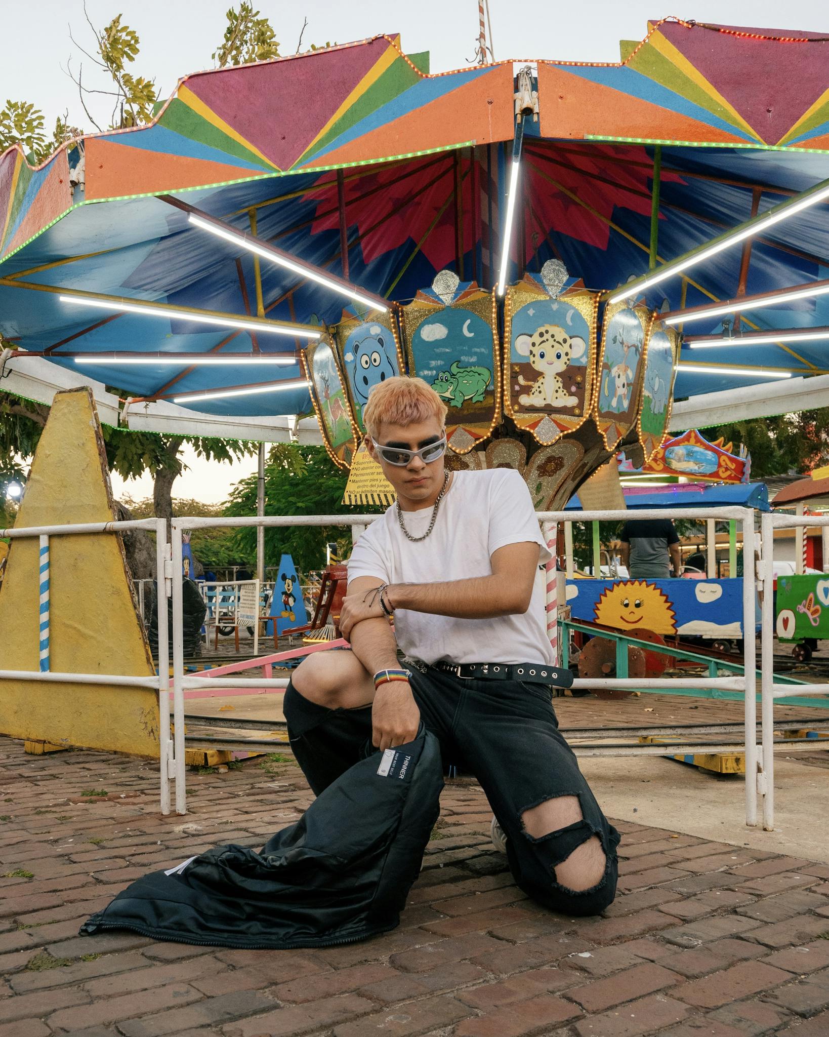A stylish young man models in front of a colorful carousel at a lively amusement park.